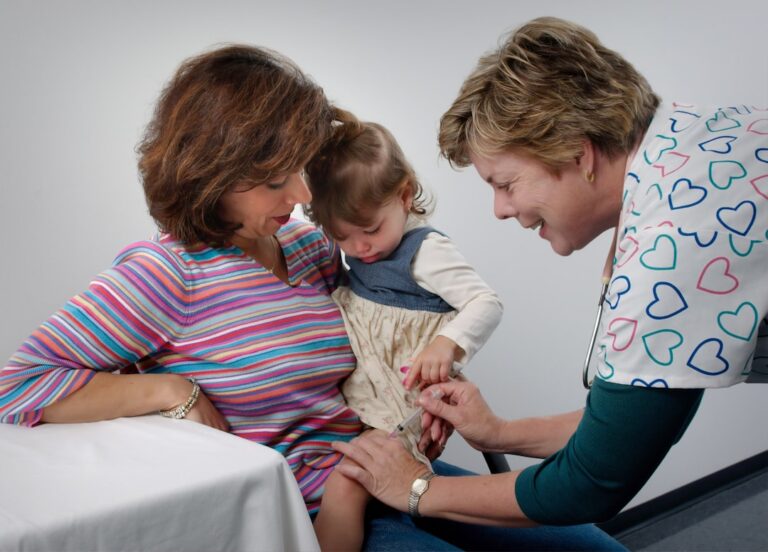 A parent measures liquid medicine with a dosing syringe for a young child sitting at a kitchen table.